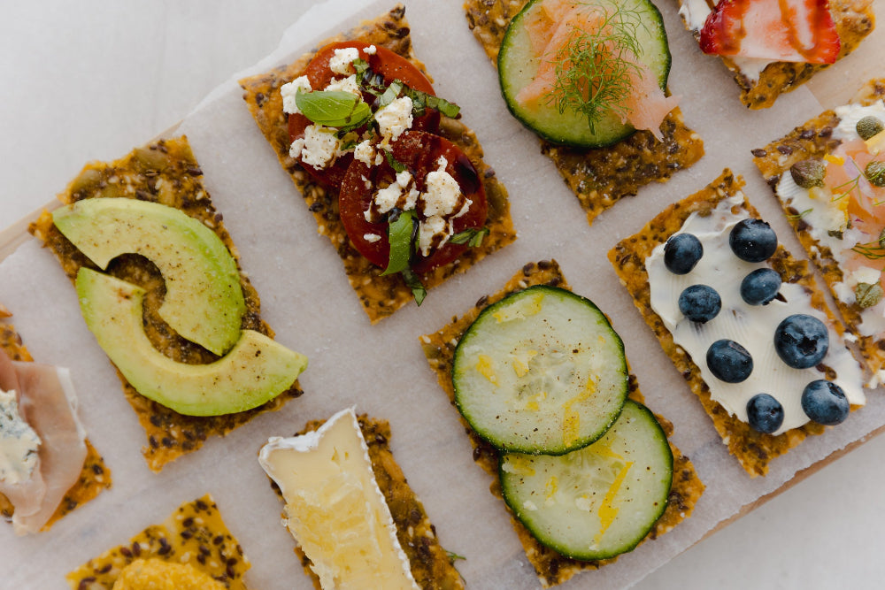 Cracker snacks with various toppings including avocado, cucumber, and blueberries on a white surface.